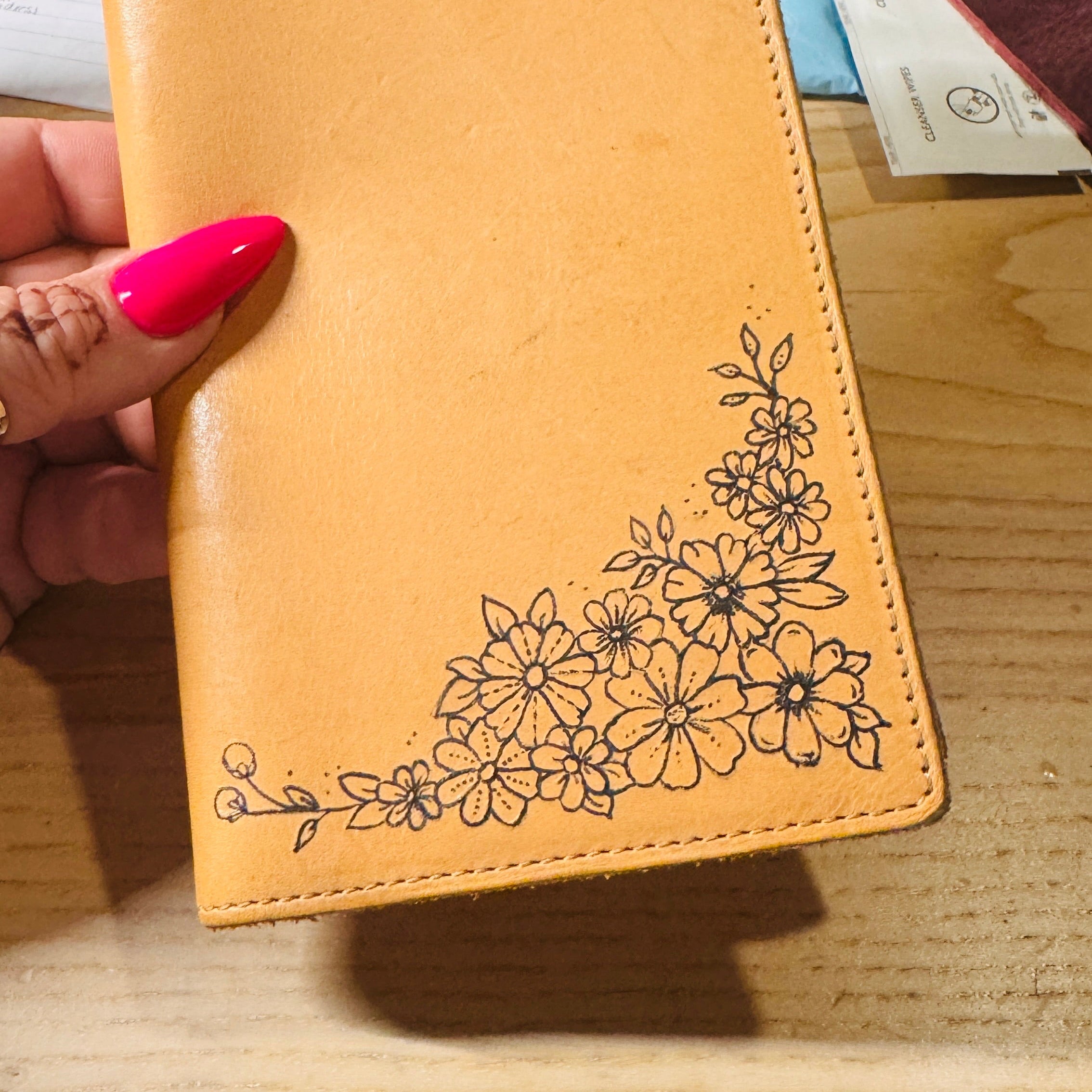 A tan leather passport cover with black floral line art in the bottom right corner, held by a hand with bright pink nail polish over a wooden desk background.